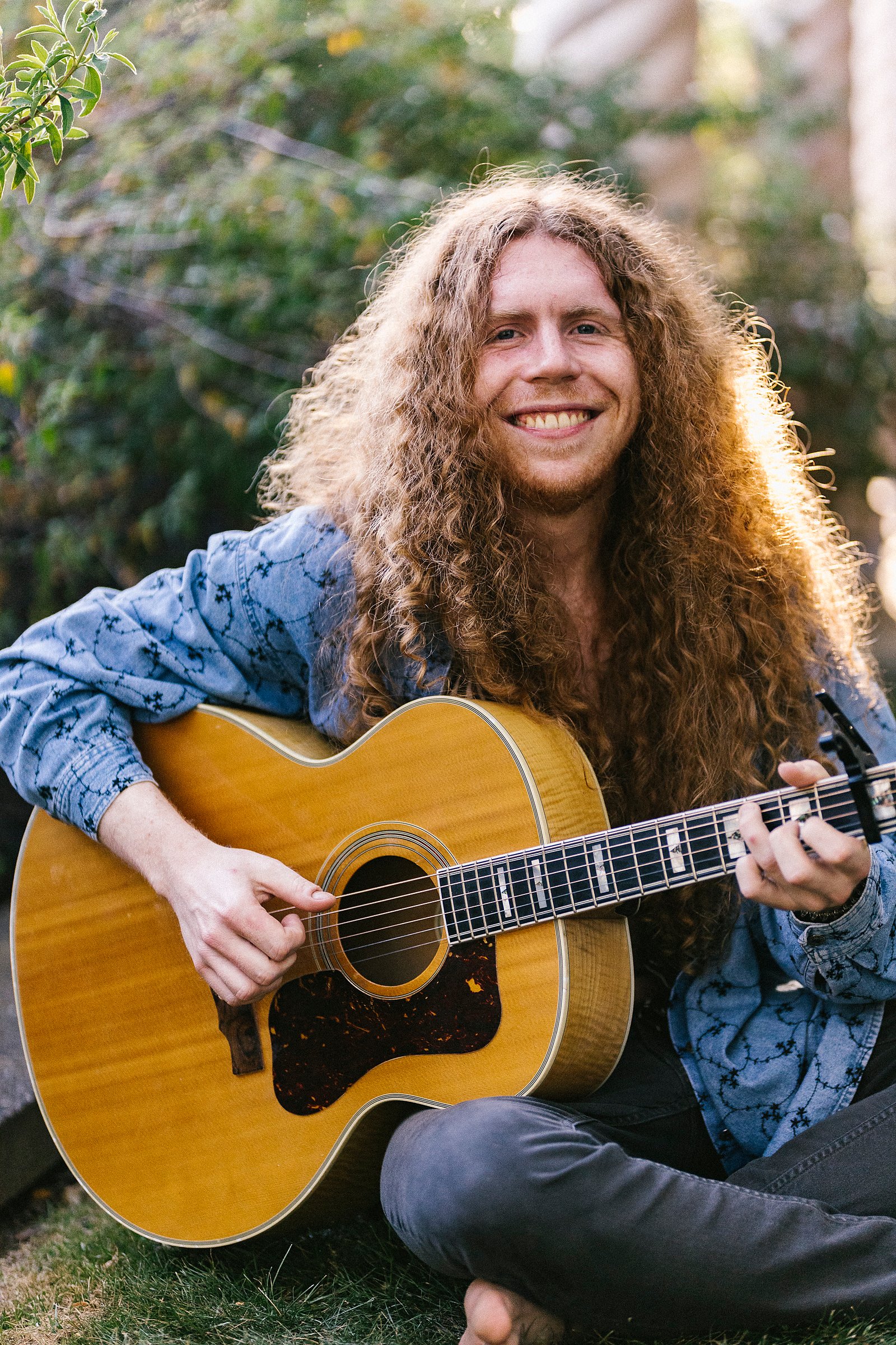 Sean Ashe smiling while seated outdoors with an acoustic guitar.