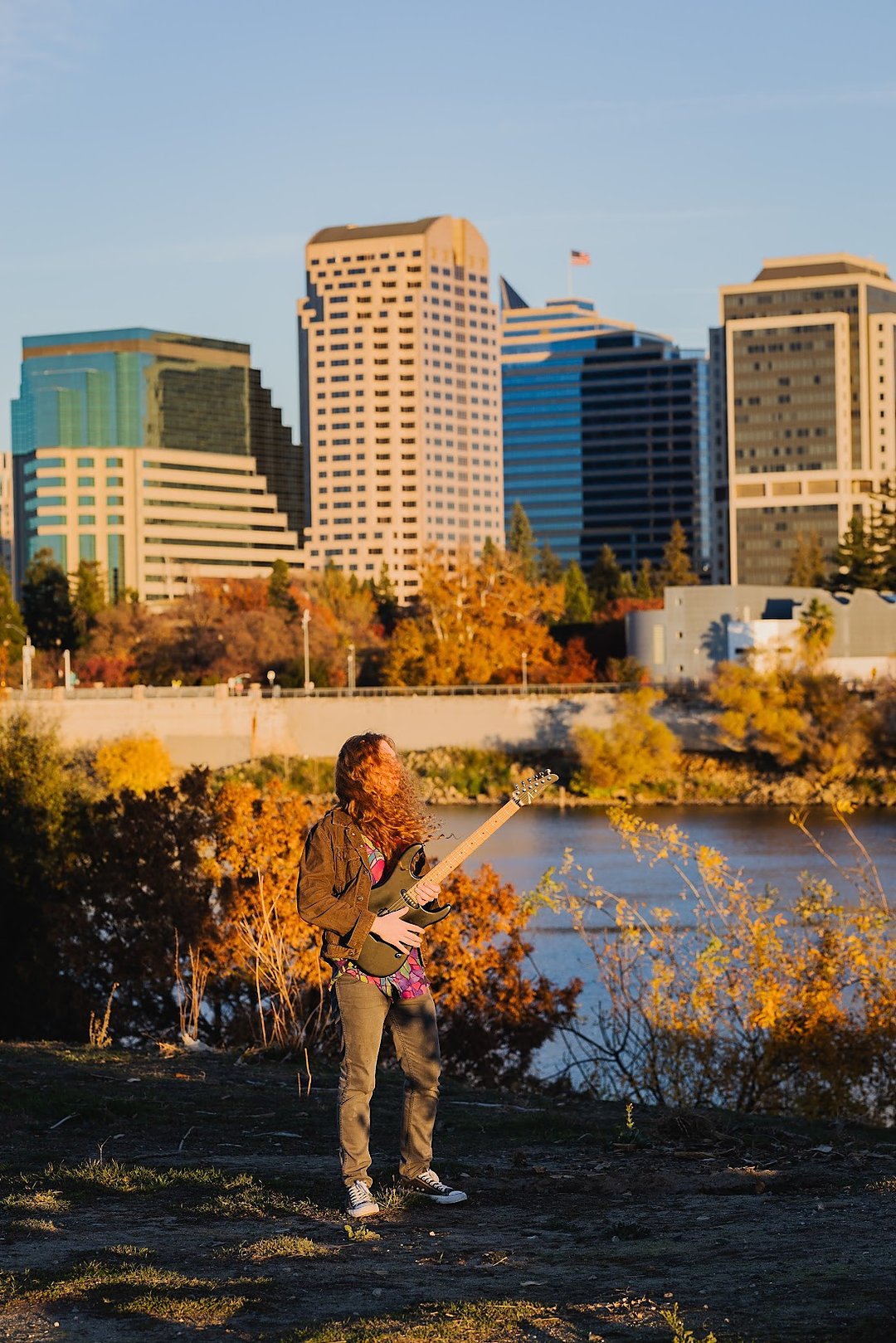 Sean Ashe playing electric guitar near the Sacramento River with the city skyline behind him.