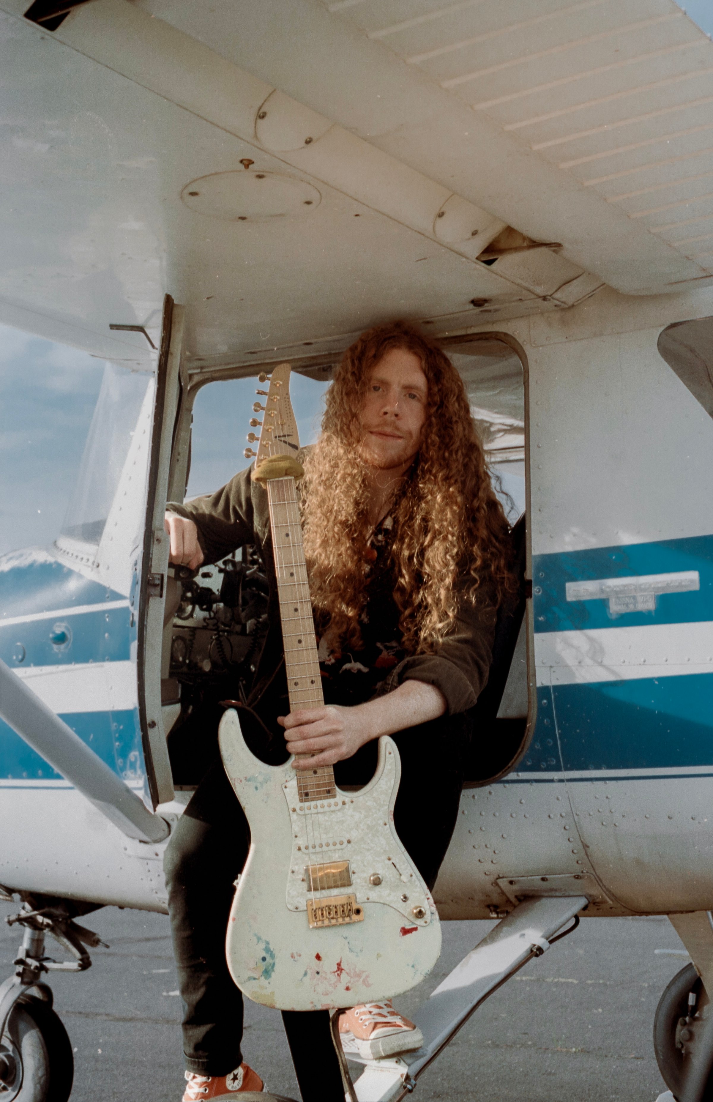 Sean Ashe seated in a small aircraft doorway holding a white electric guitar.