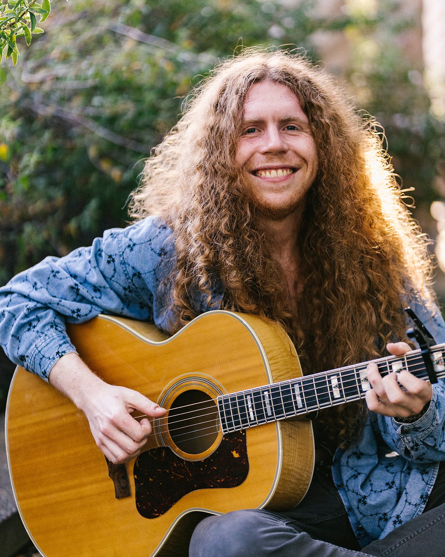 Sean Ashe smiling while seated outdoors with an acoustic guitar.