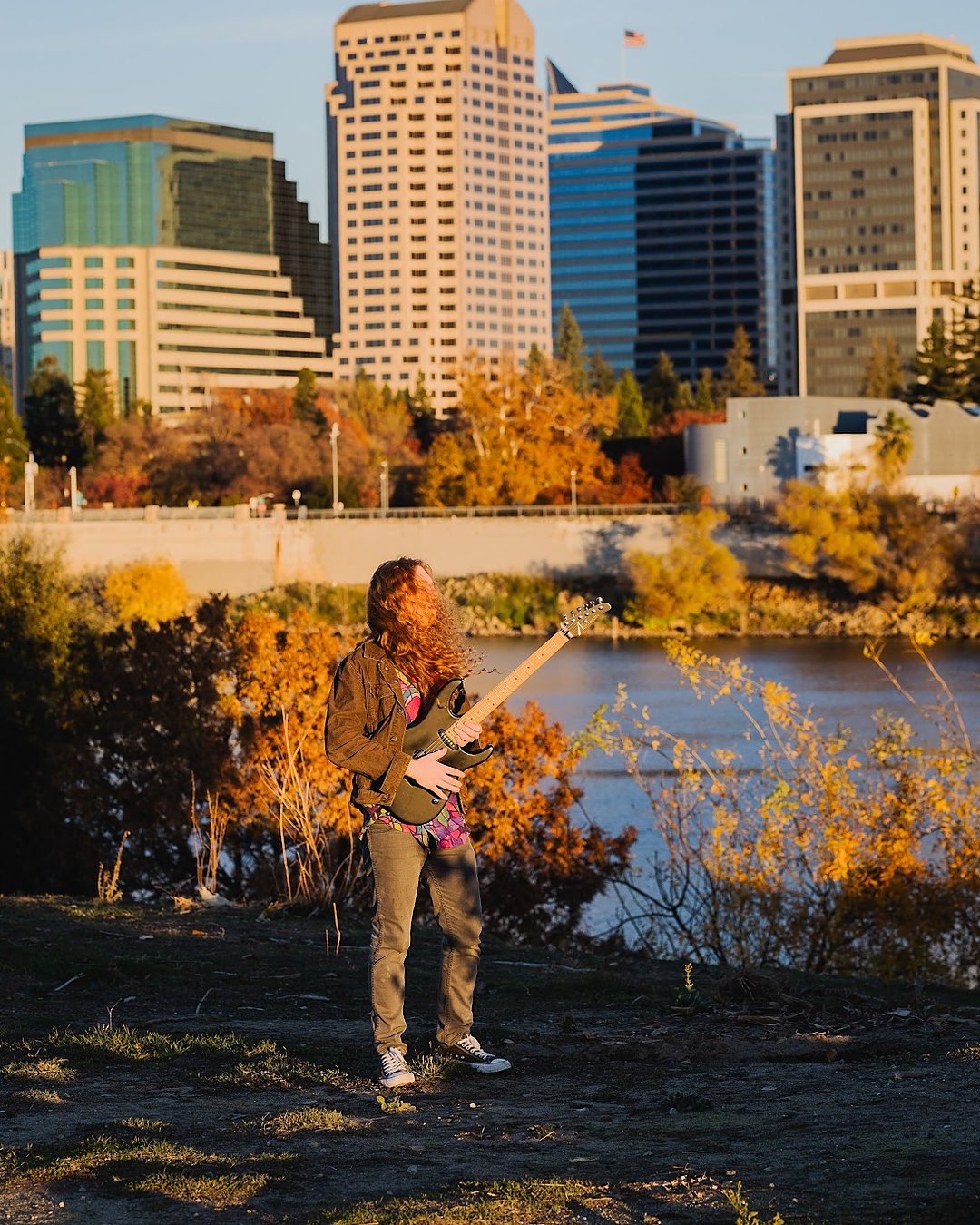Sean Ashe playing electric guitar near the Sacramento River with the city skyline behind him.