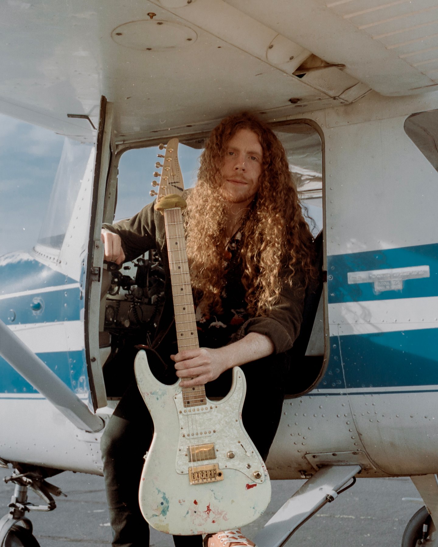 Sean Ashe seated in a small aircraft doorway holding a white electric guitar.